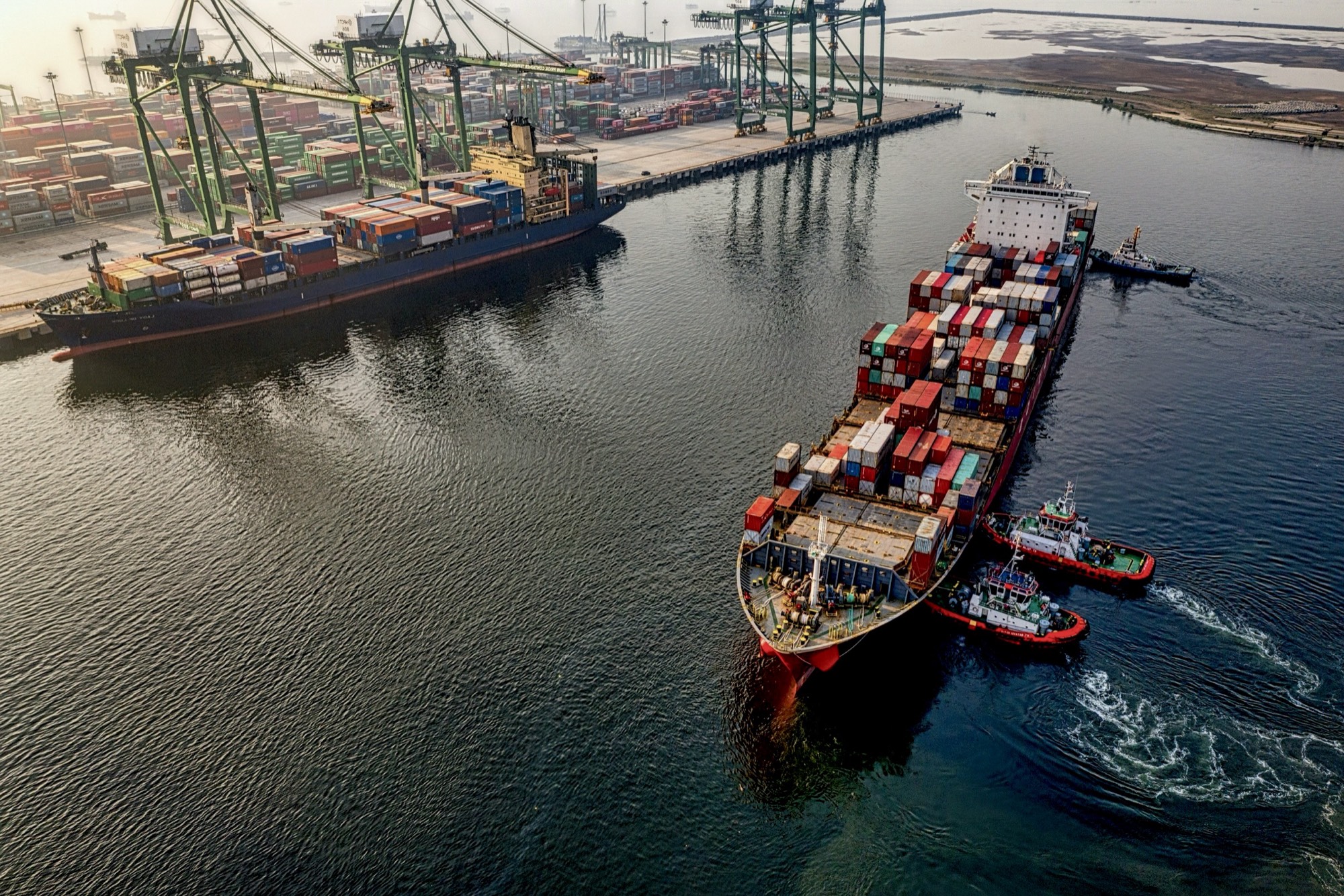 A container ship guided into port by tugboats, seen from above at sunrise
